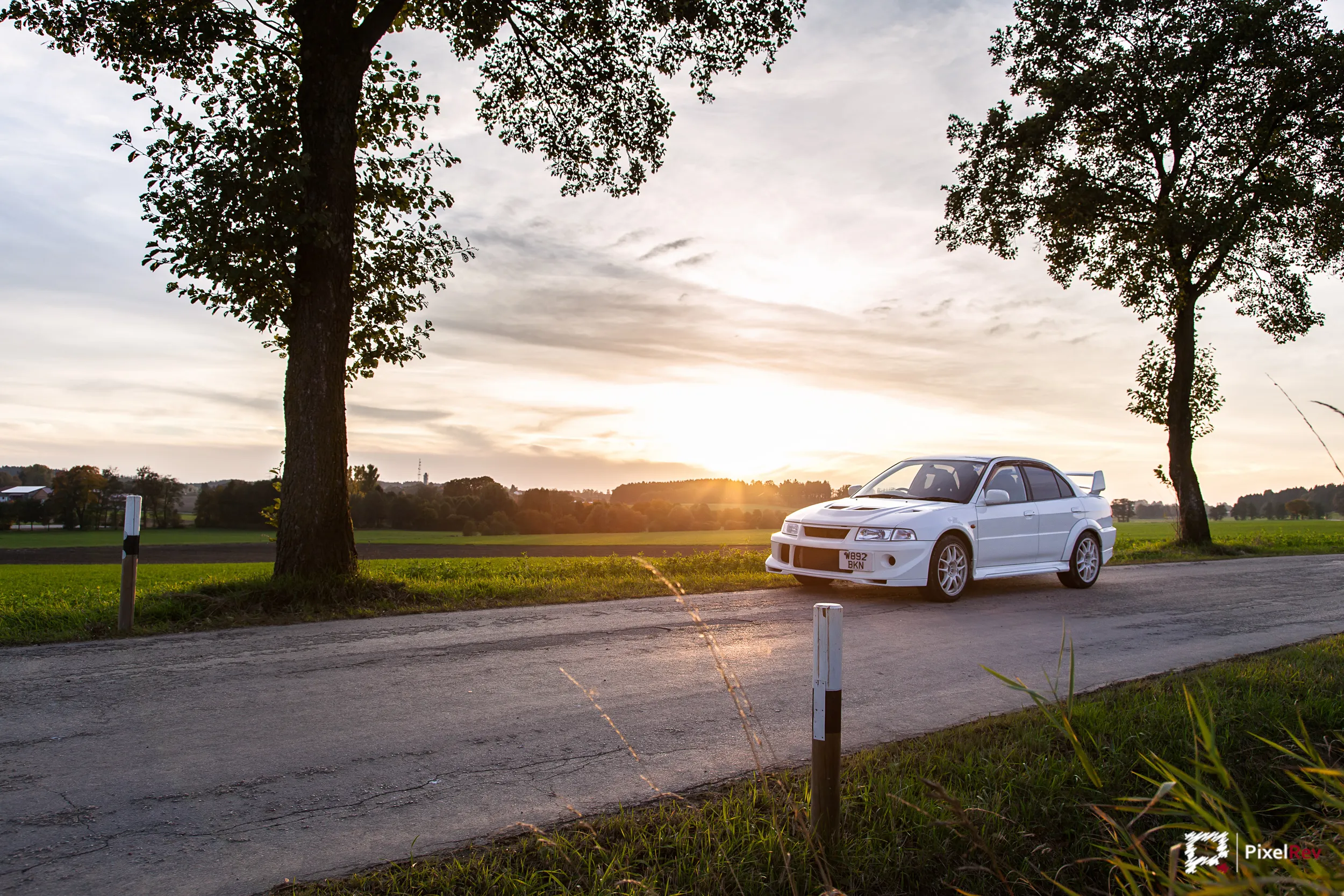 Weißer Mitsubishi Lancer Evolution VI auf einer Landstraße zwischen zwei Bäumen bei Sonnenuntergang