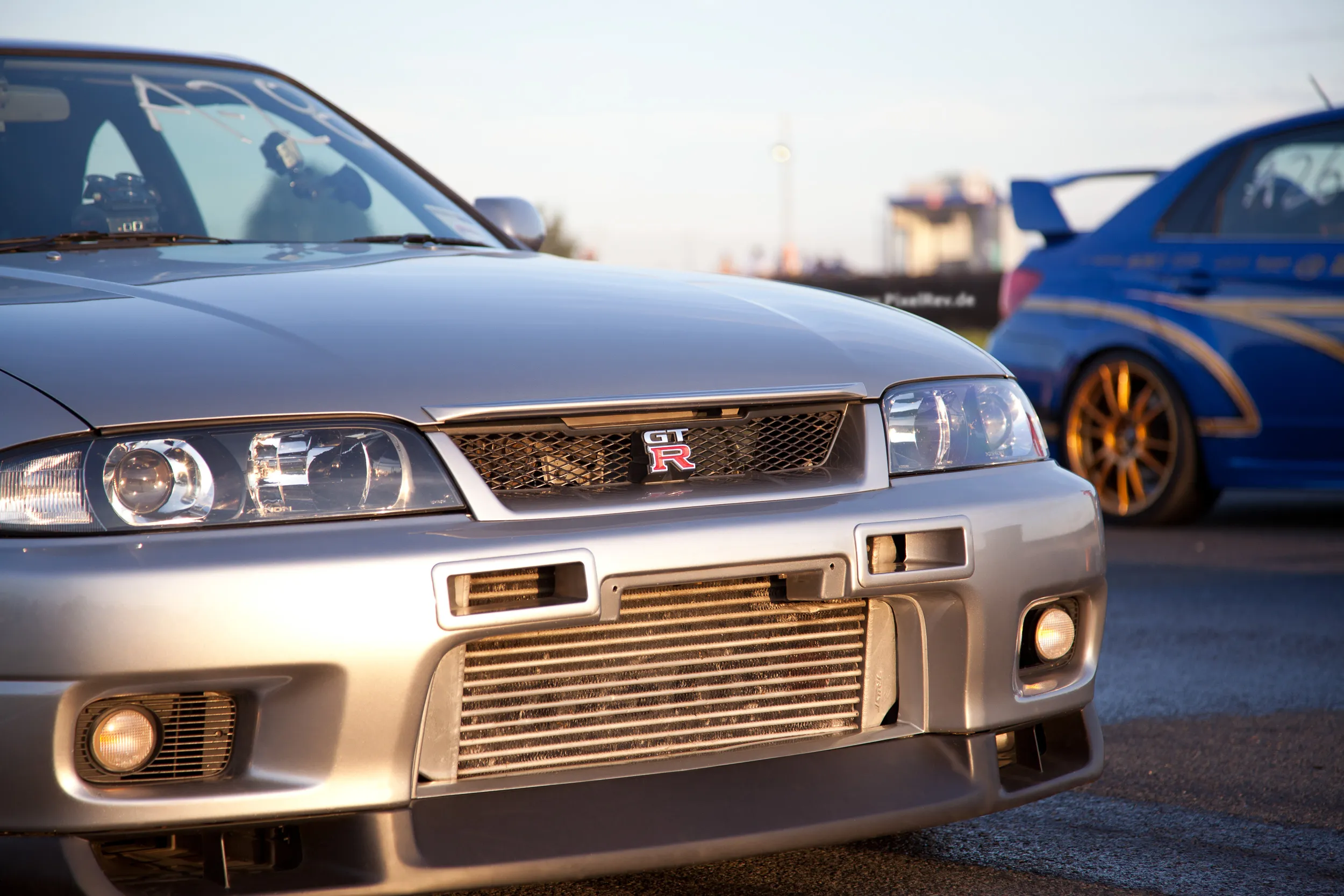Front eines silbernen Nissan Skyline R33 GT-R, daneben ein blauer Subaru Impreza