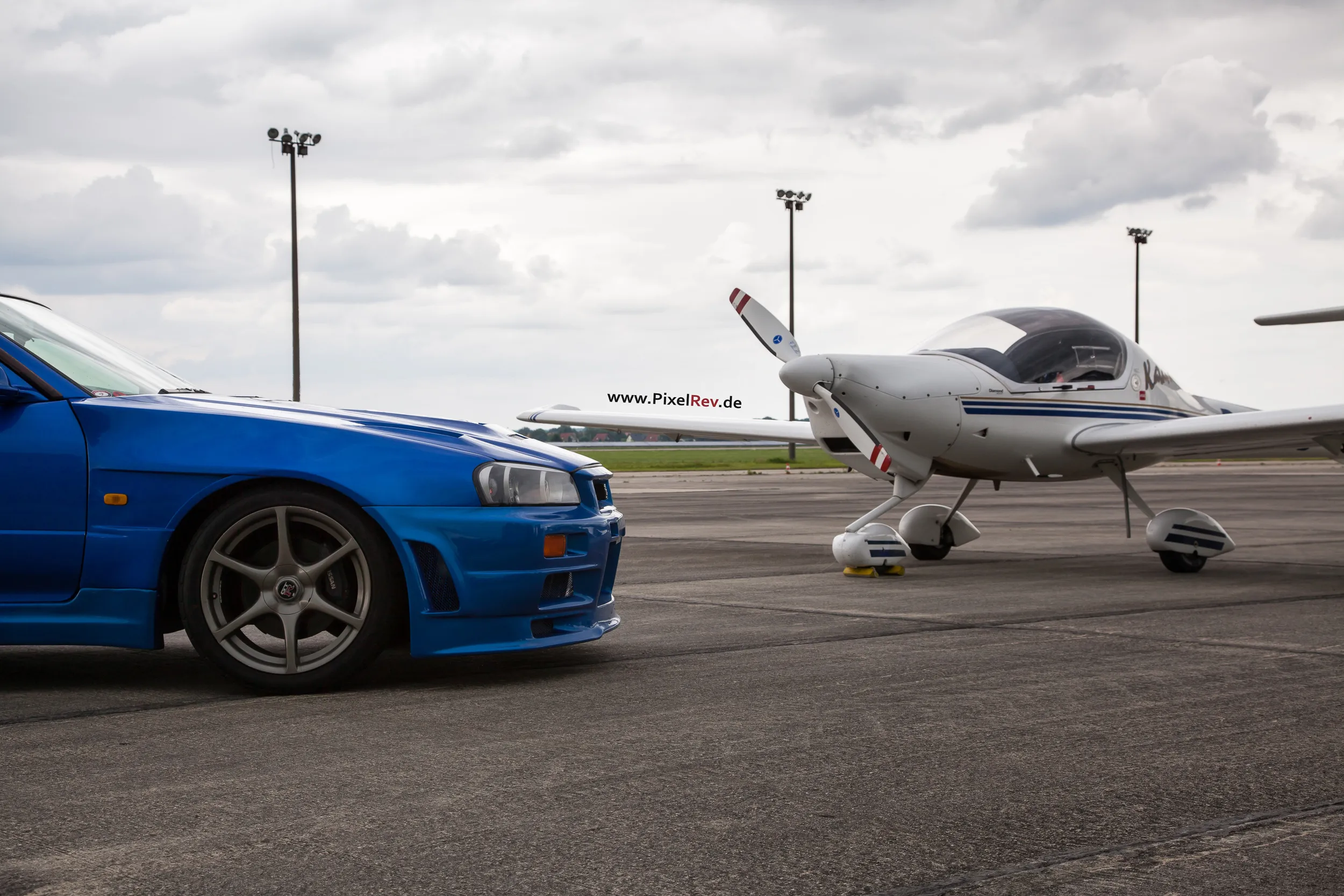 Blauer Nissan Skyline R34 GT-R neben einem Kleinflugzeug
