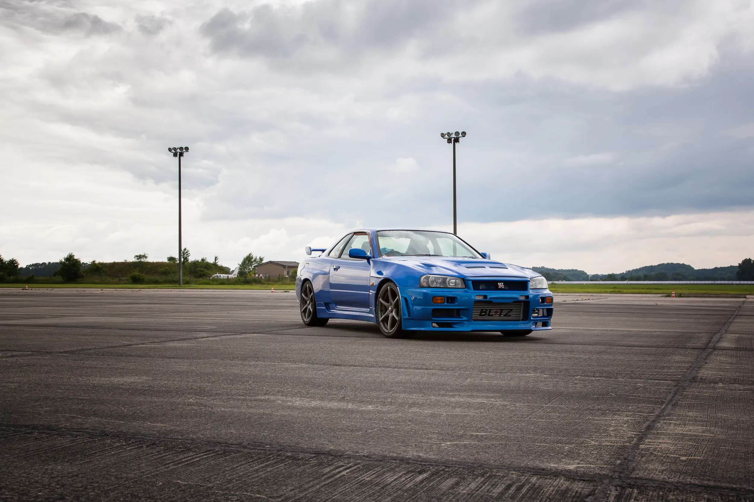 Blauer Nissan Skyline R34 GT-R auf weiter Asphaltfläche unter bewölktem Himmel