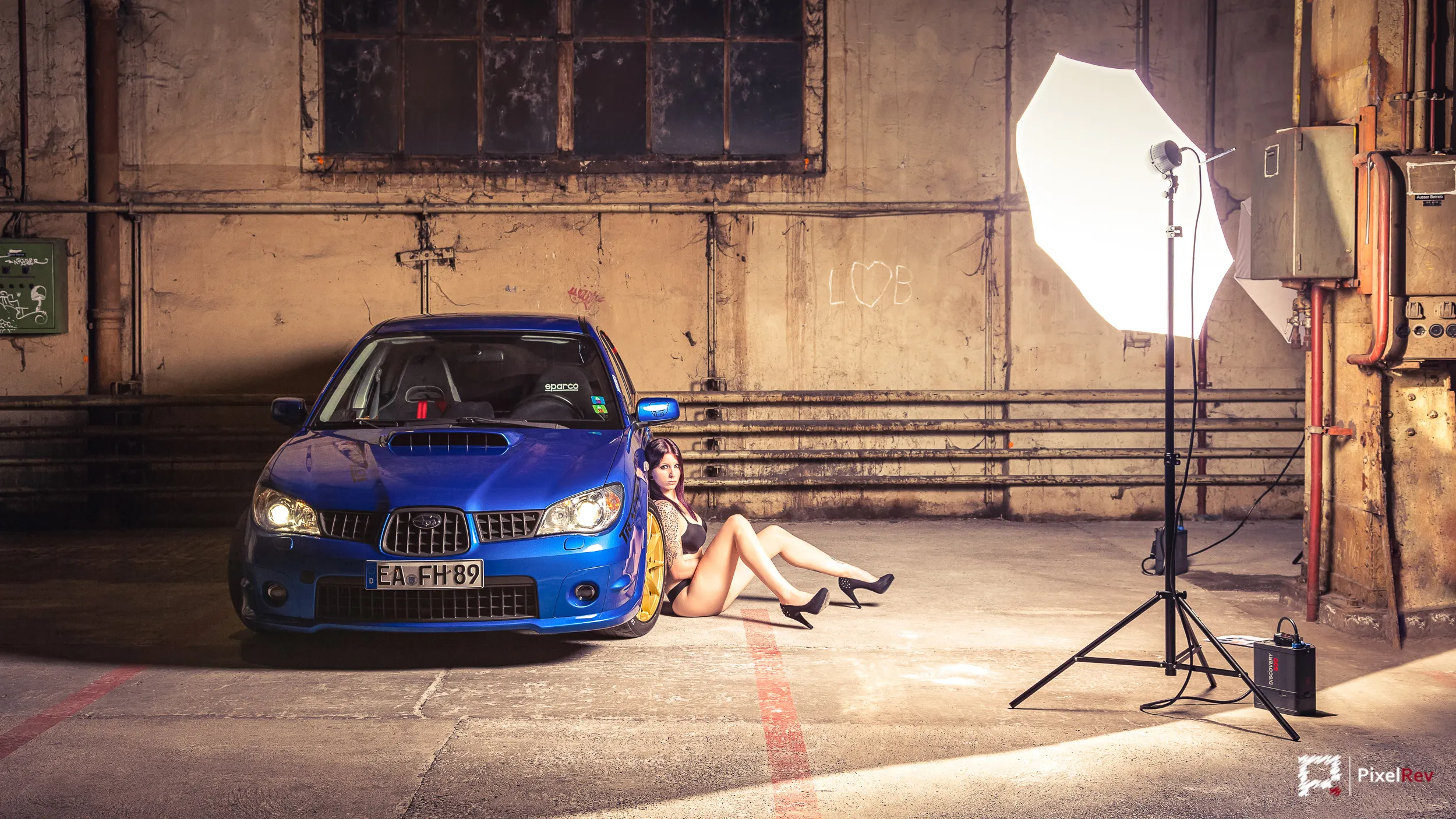 Model sitzt vor einem blauen Subaru Impreza WRX STI in einer Industriehalle, ausgeleuchtet mit Blitzschirm