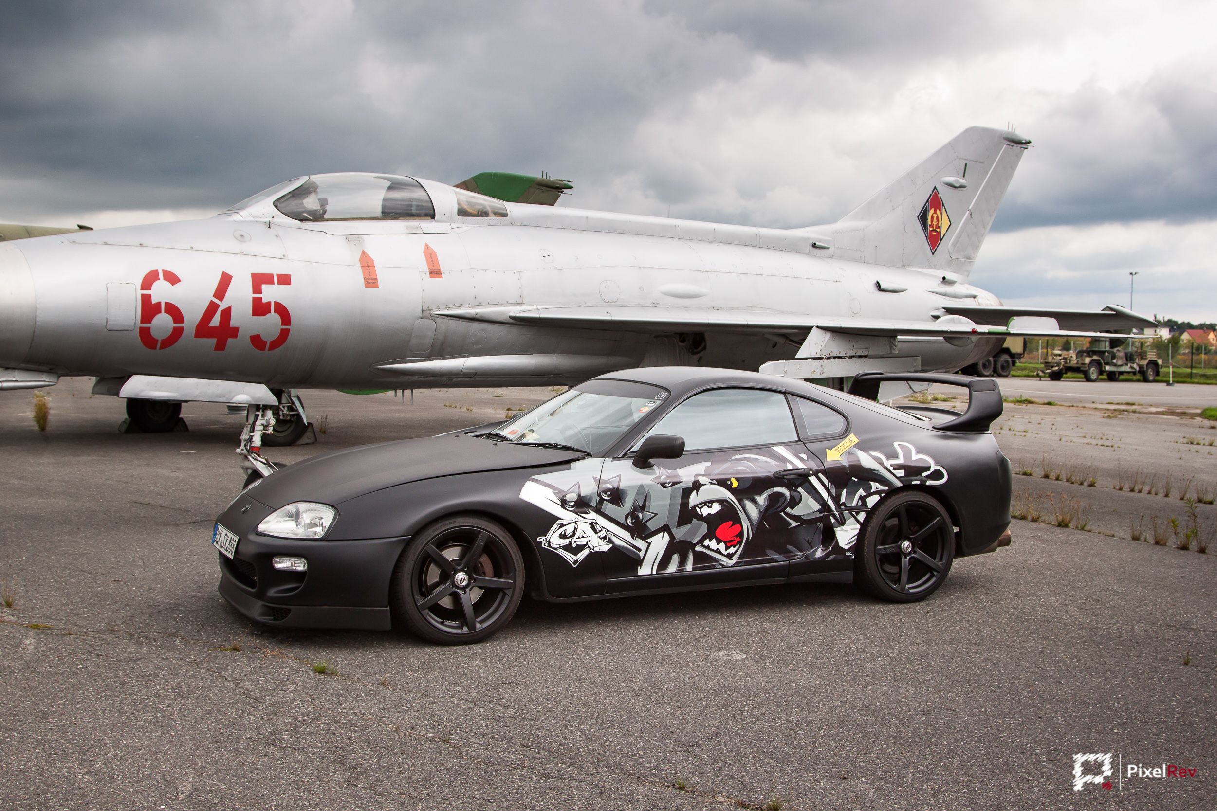 Mattschwarzer Toyota Supra mit Comic-Folierung vor einem MiG-21-Kampfjet in Berlin-Gatow