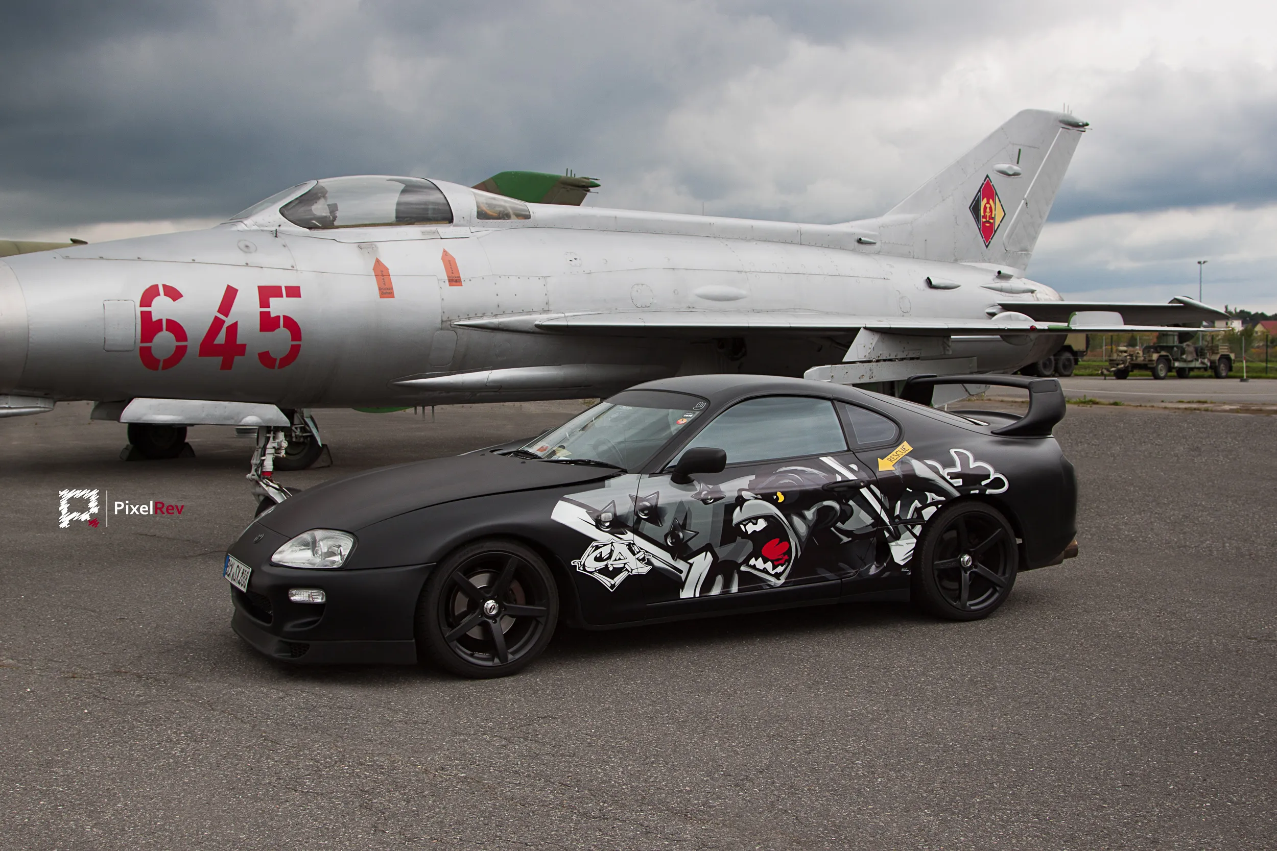 Mattschwarzer Toyota Supra mit Comic-Folierung vor einem MiG-21-Kampfjet in Berlin-Gatow