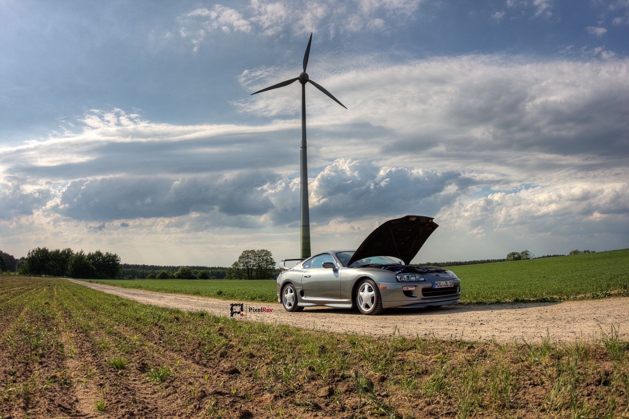 Silberner Toyota Supra Mk4 mit geöffneter Motorhaube auf einem Feldweg vor einem Windrad